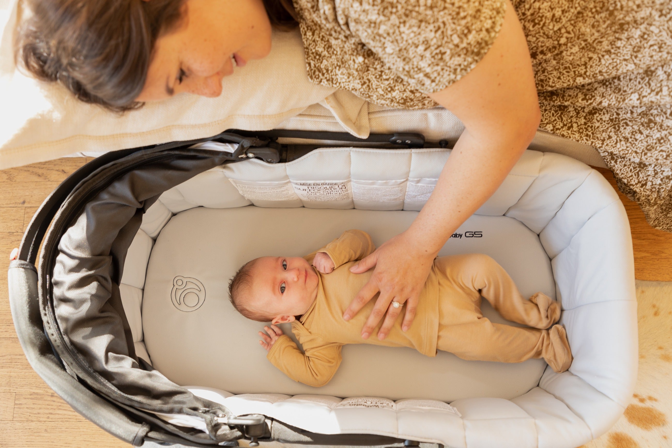 A parent leaning over the Orbit Baby G5 Travel Bassinet, resting a hand on an awake infant lying inside