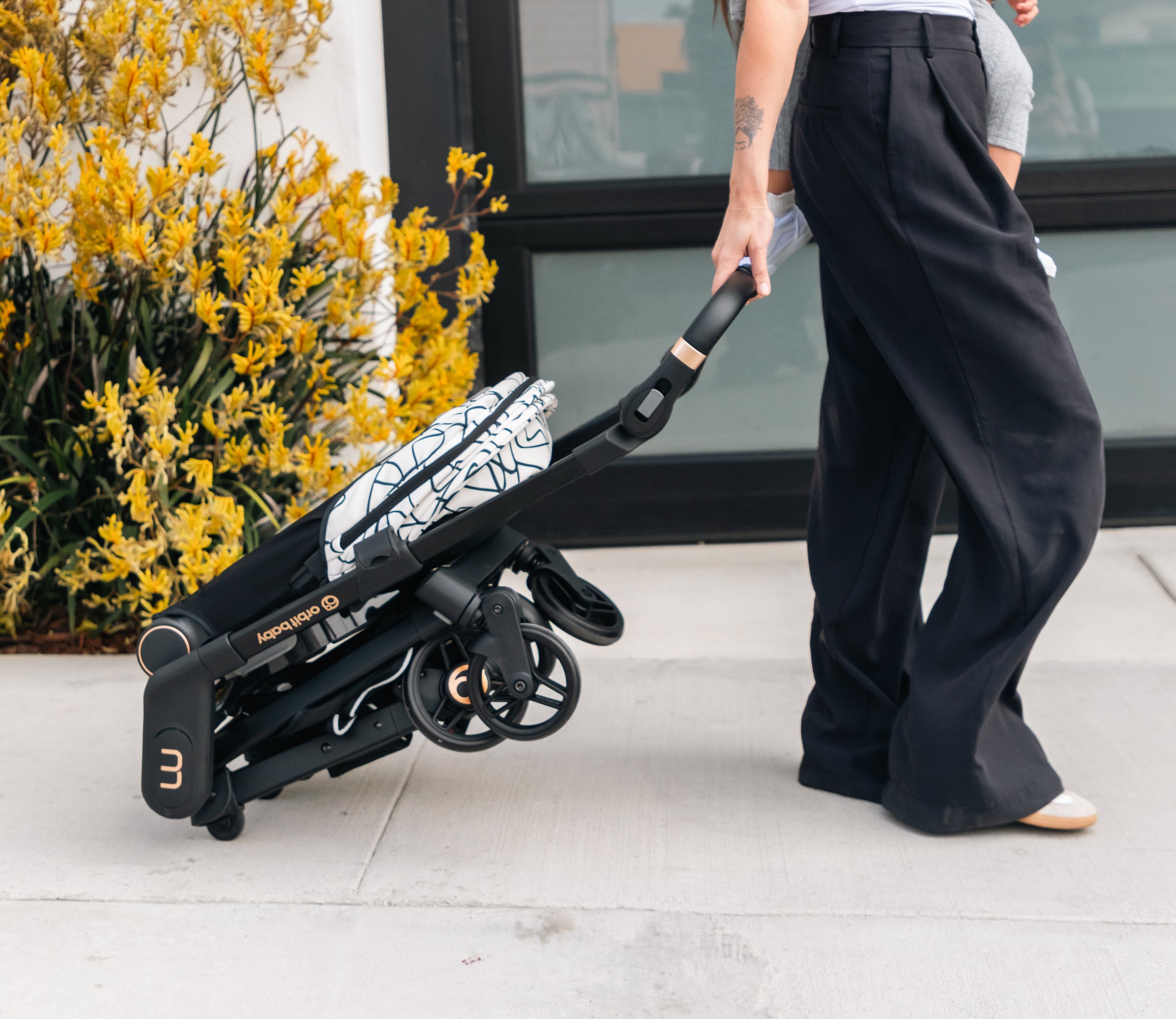 Parent rolling the Orbit Baby M+ folded into Luggage Mode by its telescoping handle along a sidewalk